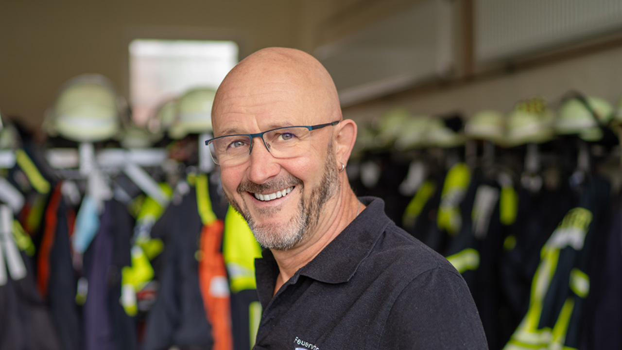 Ein lächelnder, glatzköpfiger Mann mit Brille und Bart im Porträt vor dem unscharfen Hintergrund einer Feuerwehr-Umkleide mit Schutzkleidung und Helmen.