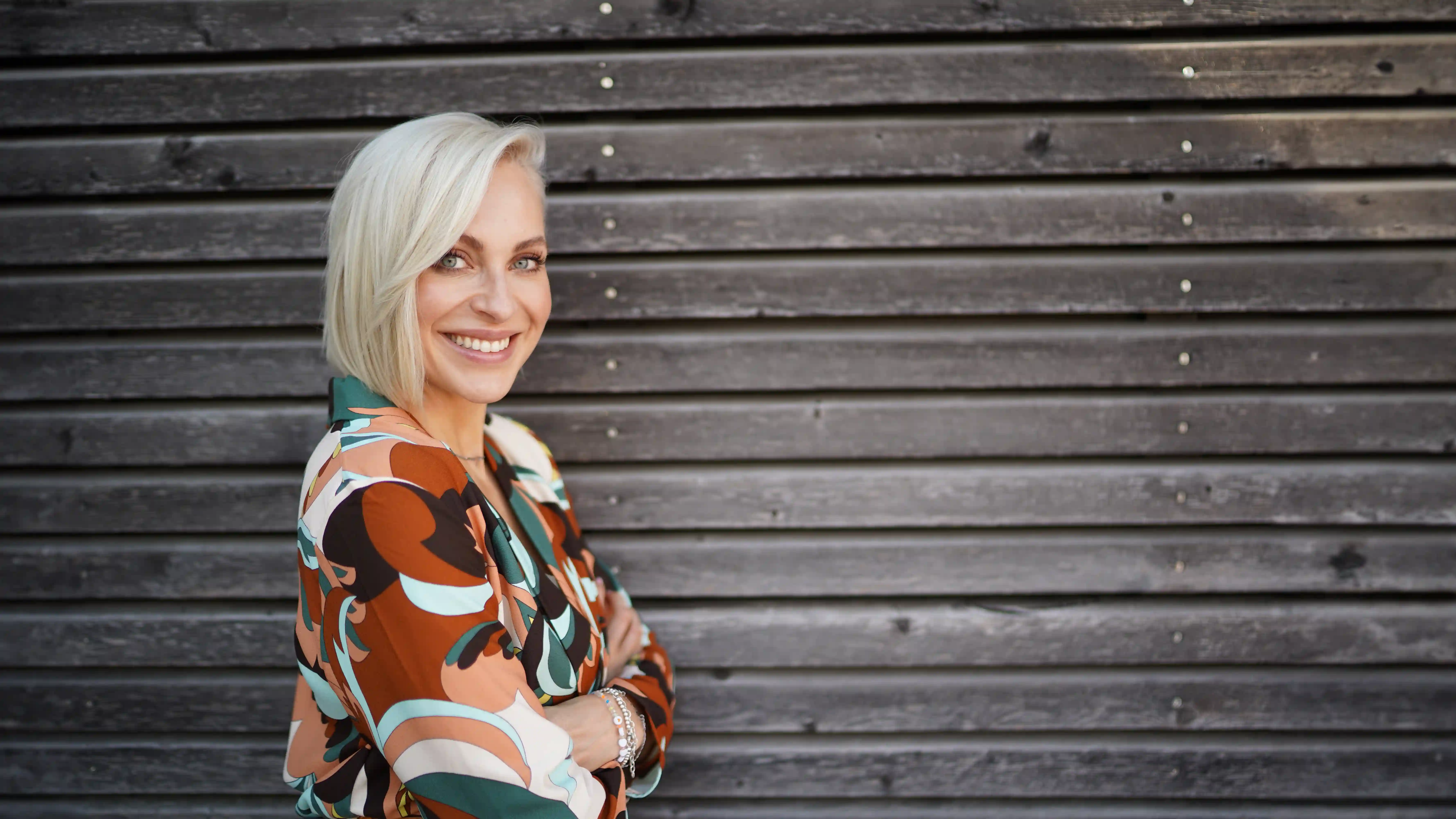 Eine lächelnde Frau mit kurzen, platinblonden Haaren im Profil vor einer grauen Holzwand. Sie trägt eine gemusterte Bluse in Orange, Braun und Grün und blickt direkt in die Kamera.
