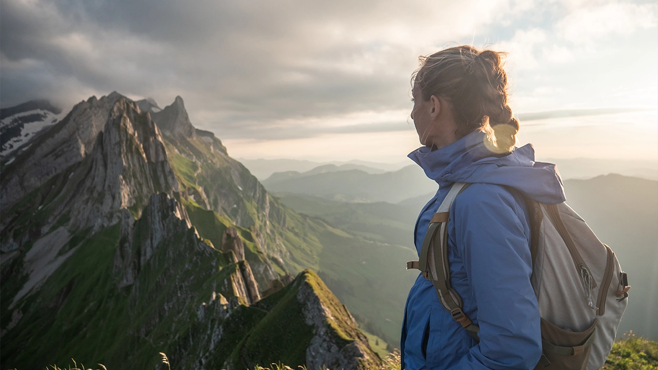 Eine Wanderin mit Rucksack und blauer Outdoor-Jacke blickt von der Seite auf eine beeindruckende Gebirgskette mit steilen, grünen Berghängen im sanften Licht des Spätnachmittags.