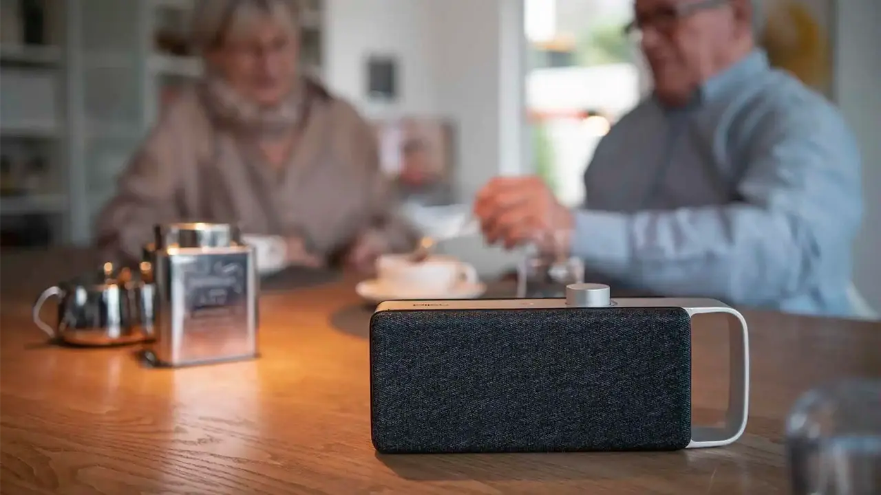 Ein moderner, tragbarer Lautsprecher mit dunkelgrauem Stoffbezug und silbernem Tragegriff steht im Fokus auf einem Holztisch. Im unscharfen Hintergrund sitzt ein Paar beim Kaffeetrinken.