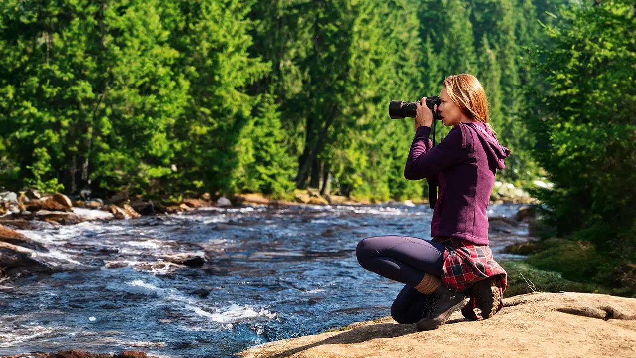 Eine Frau in Wanderbekleidung hockt am felsigen Ufer eines wilden Flusses und fotografiert die Landschaft mit einer Kamera. Im Hintergrund erstreckt sich ein dichter, grüner Nadelwald.
