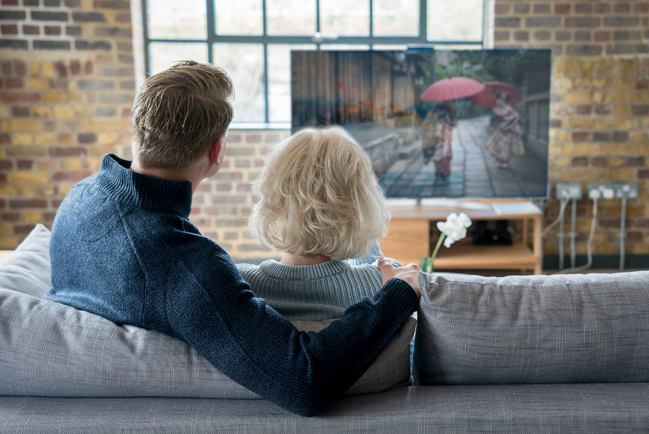 Ein Paar sitzt von hinten gesehen auf einem Sofa und schaut gemeinsam auf einen großen Fernseher. Auf dem Bildschirm läuft eine Szene mit traditionell gekleideten japanischen Frauen mit roten Regenschirmen.