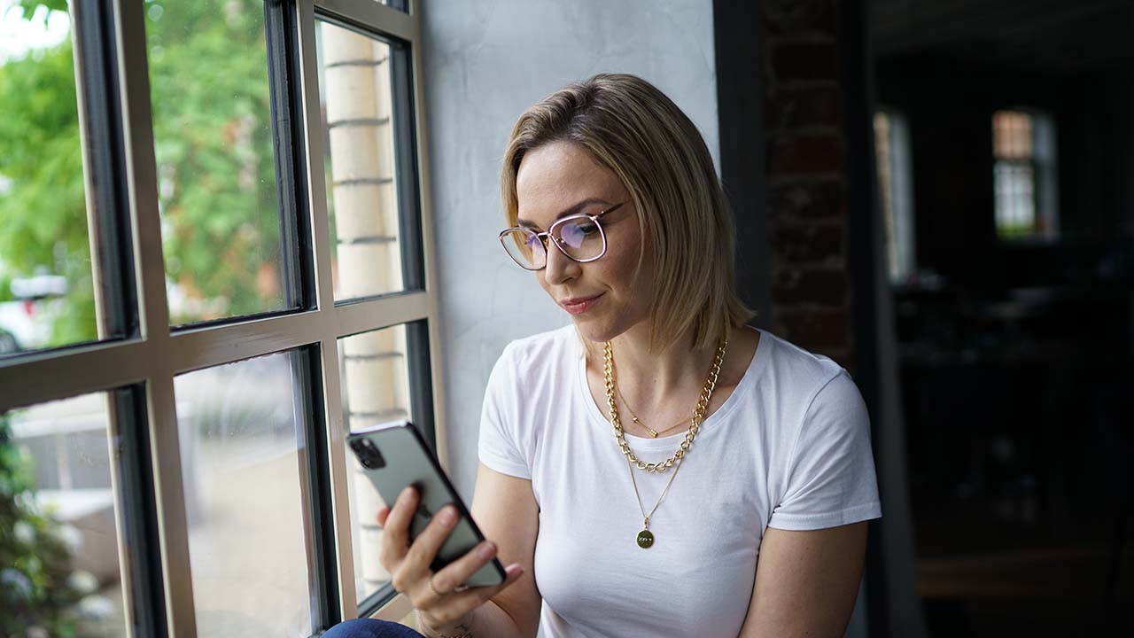Eine Frau sitzt vor einem Fenster und schaut dabei auf ihr Handy, sie trägt eine Blaulichtfilterbrille