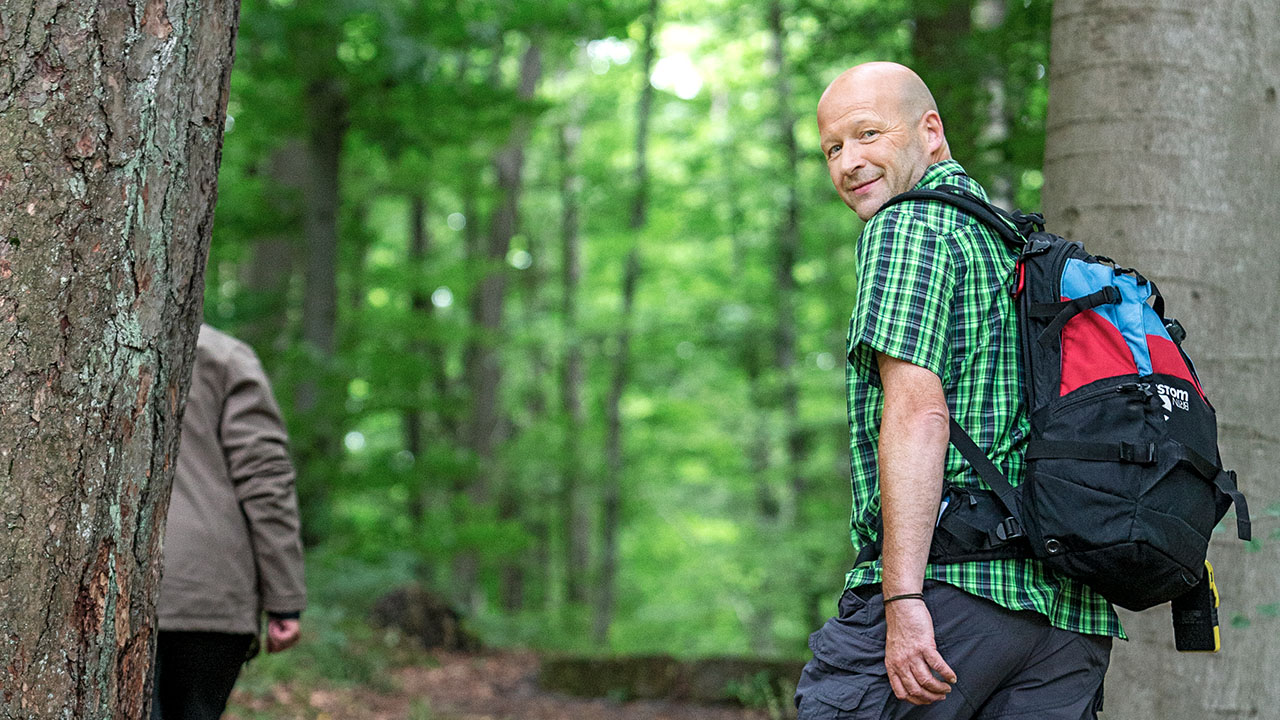 Ein lächelnder, glatzköpfiger Wanderer mit Rucksack und grün-schwarz kariertem Hemd blickt im Profil über die Schulter zurück in die Kamera. Er befindet sich auf einem Pfad inmitten eines dichten, grünen Waldes.