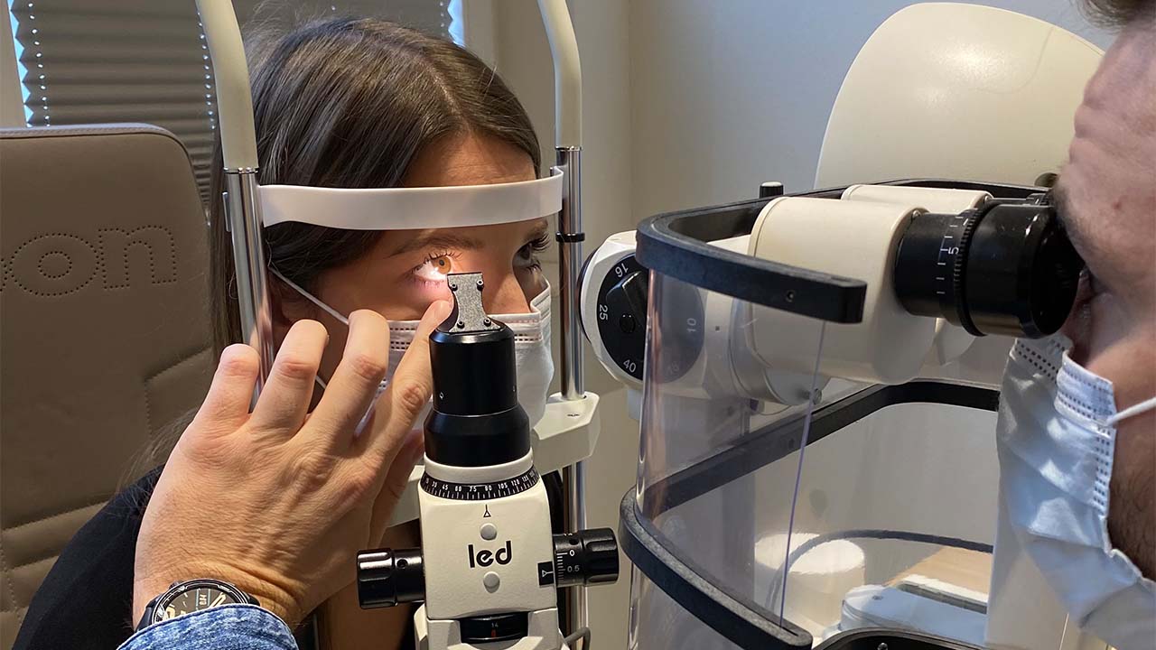 Eine medizinische Augenuntersuchung beim Optiker oder Augenarzt: Das Auge einer Patientin mit Atemschutzmaske wird mithilfe einer Spaltlampe vergrößert und durchleuchtet.
