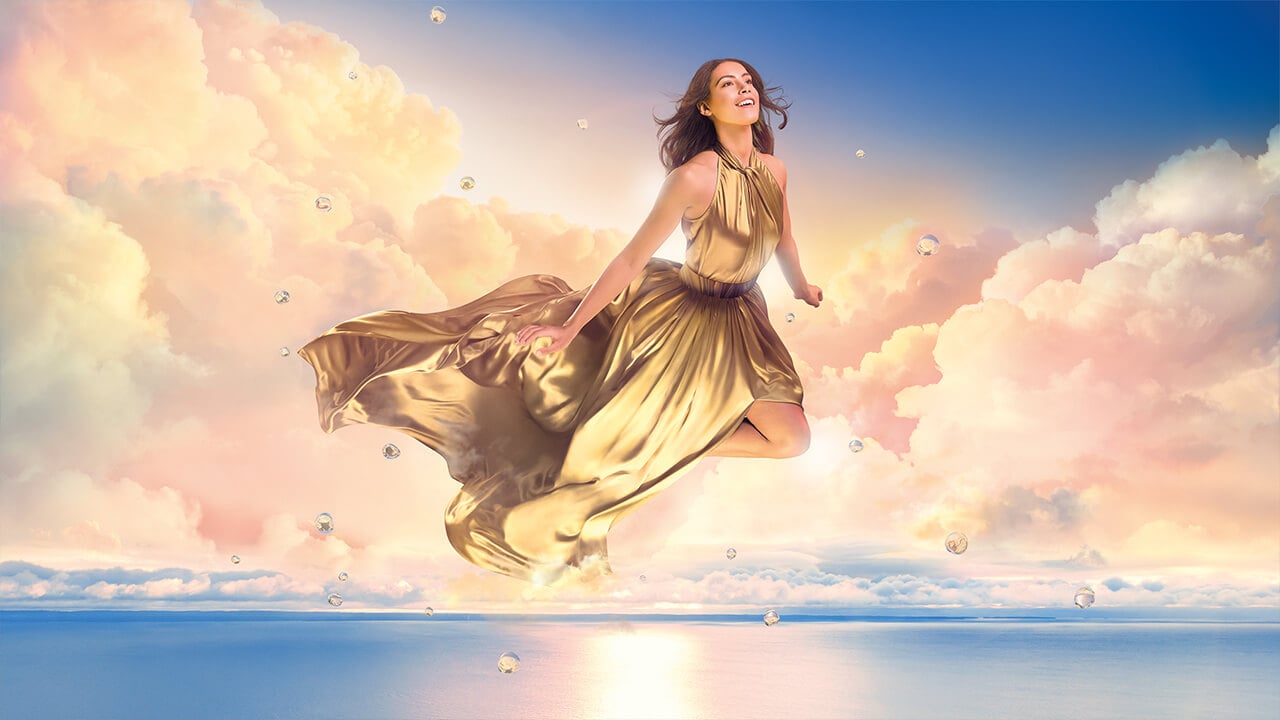 Visualisierung des Tragekomforts: Eine lächelnde Frau in einem goldenen, fließenden Kleid schwebt unbeschwert durch weiche, gold-pinkfarbene Wolken, umgeben von schwebenden Wassertropfen und Seifenblasen.
